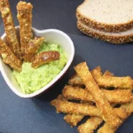 sobre una pizarra varios palitos de pan crujiente rebozados de queso, unas rebanadas de pan, y un cuenco en forma de corazón donde varios palitos están insertados en salsa de aguacate