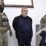 Former Karabakh separatist leader Ruber Vardanyan is shown handcuffed with his hands behind his back next to two Azerbaijani soldiers; an old picture of him when he was in power is displayed in one corner.