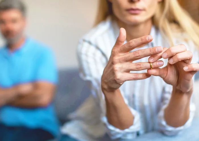 un matrimonio con una mujer joven y rubia en primer plano quitándose el anillo de casados, y de fondo el marido mirándola