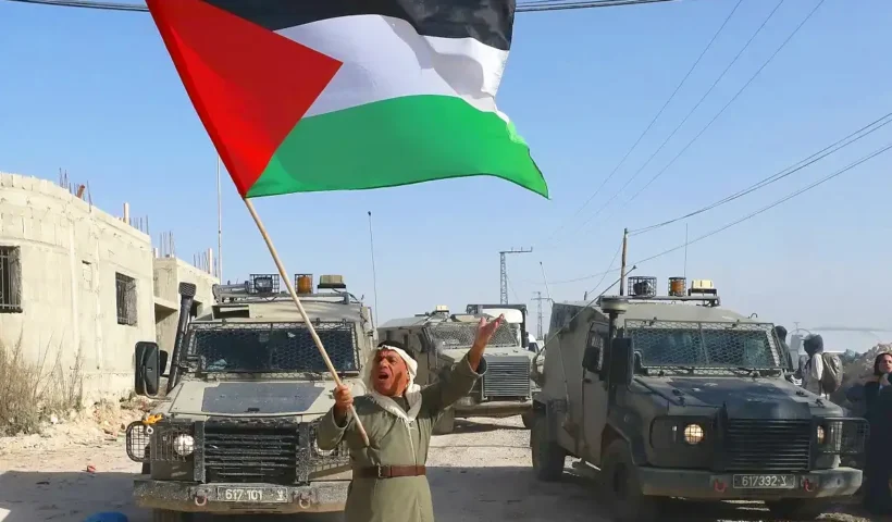 un palestino ondea una gran bandera palestina frente un convoy de fuerzas israelíes en la Cisjordania ocupada