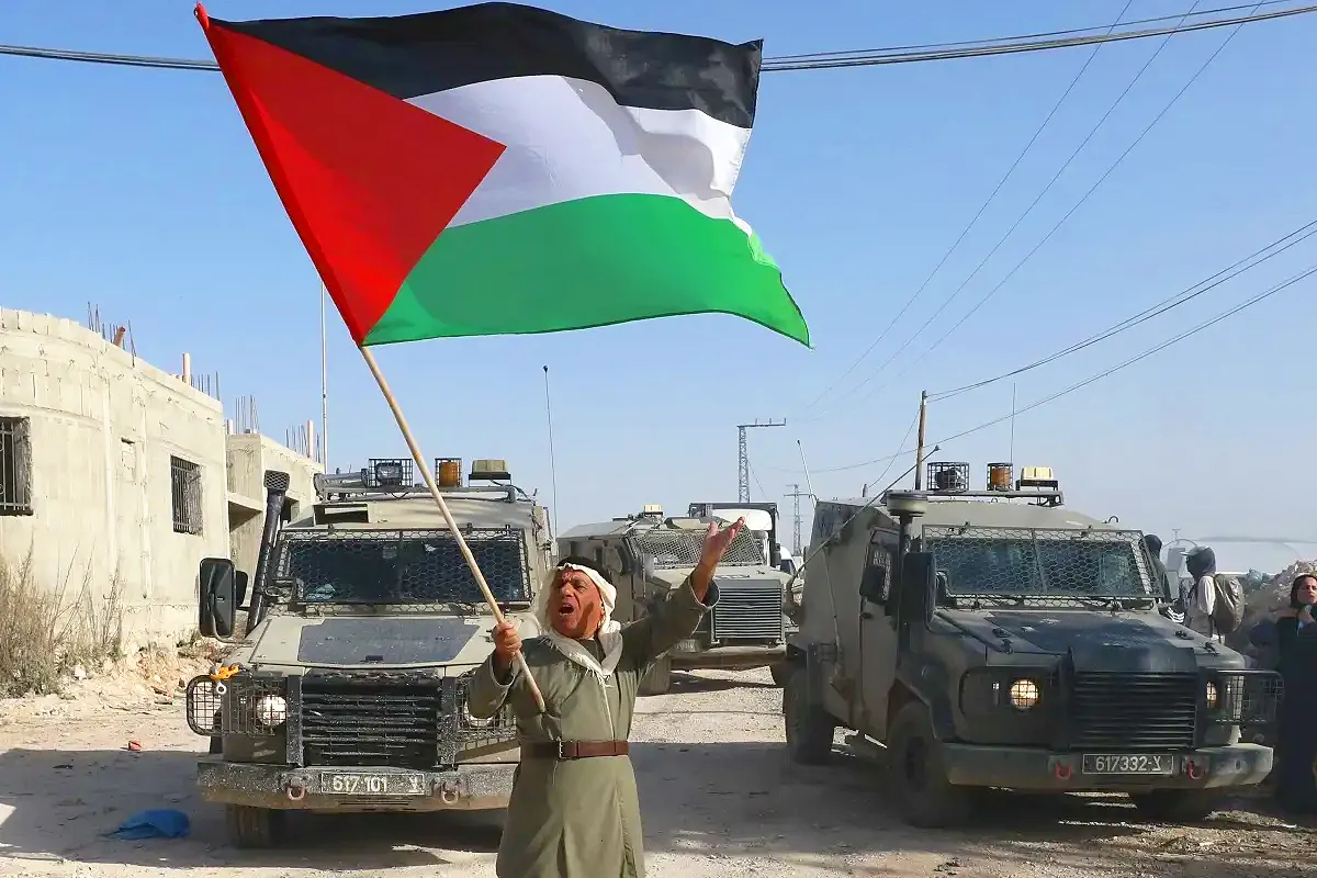 un palestino ondea una gran bandera palestina frente un convoy de fuerzas israelíes en la Cisjordania ocupada