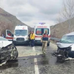dos coches destrozados tras haber chocado frontalmente en una carretera, ambos cara a cara y uno de ellos montado sobre una valla; en medio hay dos ambulancias y personal de seguridad
