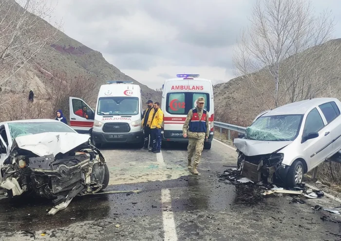 dos coches destrozados tras haber chocado frontalmente en una carretera, ambos cara a cara y uno de ellos montado sobre una valla; en medio hay dos ambulancias y personal de seguridad