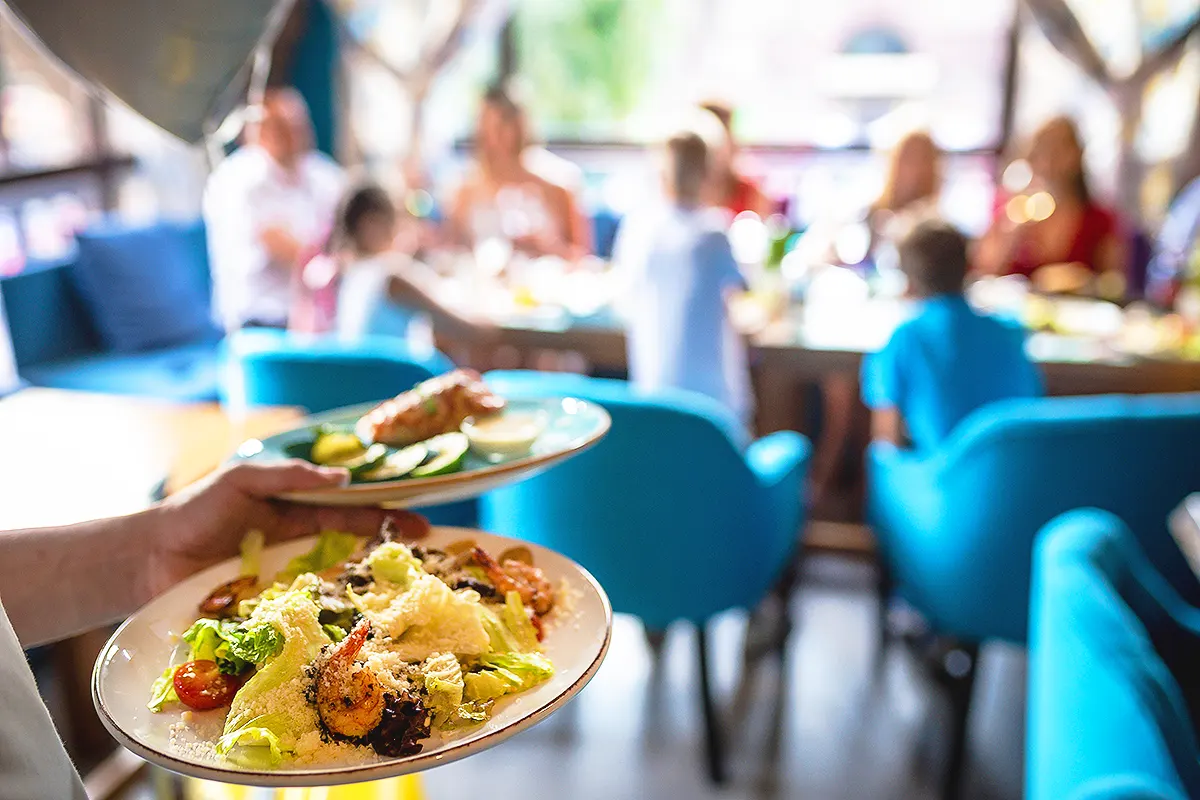 un camarero sirviendo dos platos de comida con ensalada y marisco a una mesa donde hay una familia con varios niños