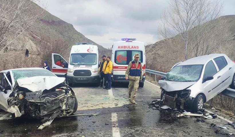 two cars wrecked after colliding head-on on a road, both face each other and one of them mounted on a fence; in between are two ambulances and security personnel