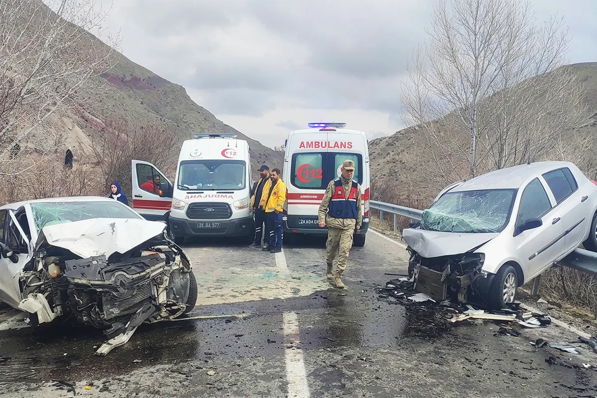 two cars wrecked after colliding head-on on a road, both face each other and one of them mounted on a fence; in between are two ambulances and security personnel
