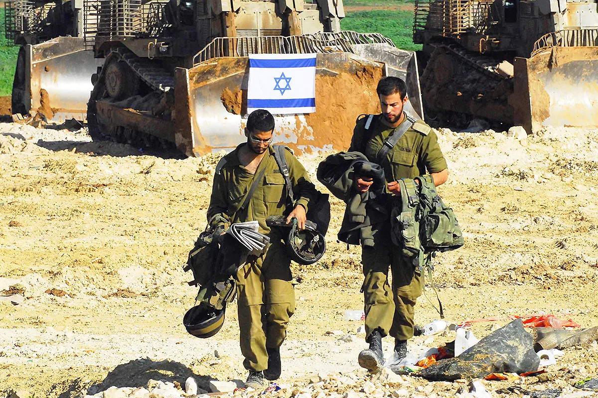 Two Israeli soldiers walk with some of their equipment removed, behind them two bulldozers and an Israeli flag.