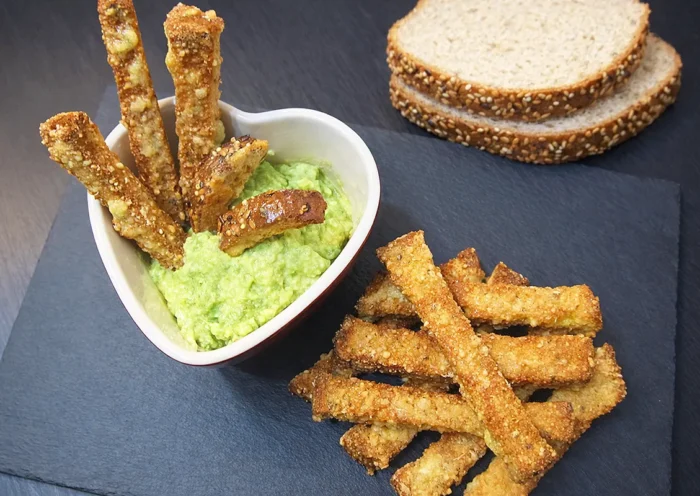 on a blackboard several cheese coated crispbread sticks, some slices of bread, and a heart-shaped bowl where several sticks are dipped in avocado sauce