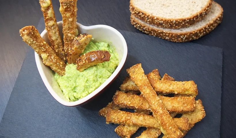 on a blackboard several cheese coated crispbread sticks, some slices of bread, and a heart-shaped bowl where several sticks are dipped in avocado sauce