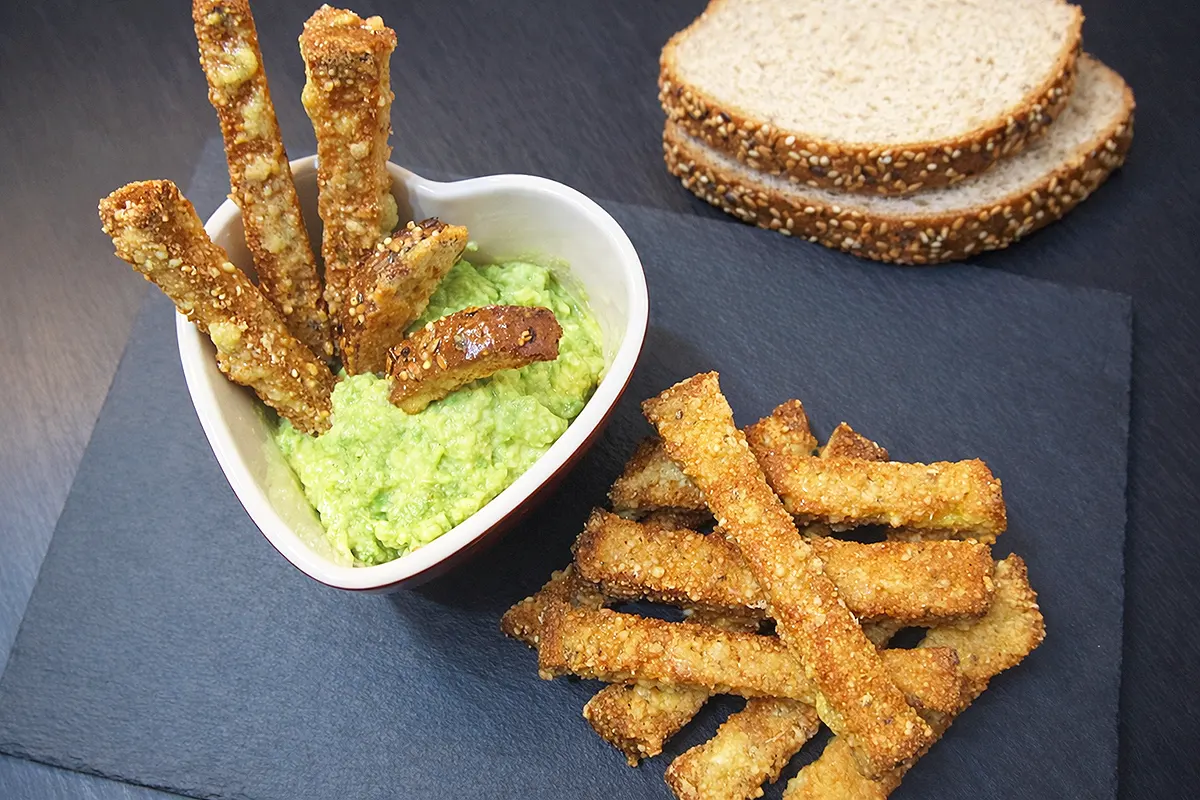 on a blackboard several cheese coated crispbread sticks, some slices of bread, and a heart-shaped bowl where several sticks are dipped in avocado sauce