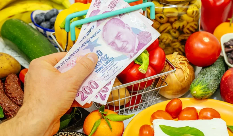 varias frutas, verduras y otros alimentos de fondo, y sobre ellos una mano mostrando un pequeño carrito de la compra y sosteniendo varios billetes de liras turcas