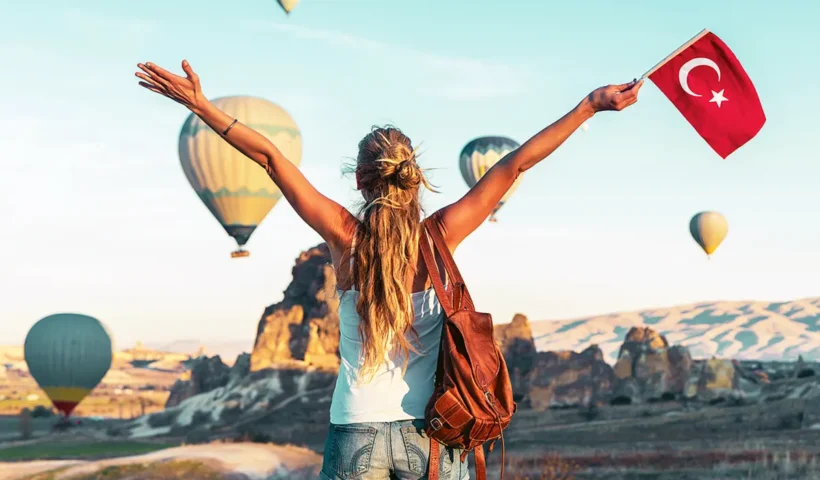 una mujer vestida con camiseta y pantalones cortos y situada de espaldas ondea una bandera turca mientras contempla alzarse los globos aerostáticos sobre el paisaje de Capadocia