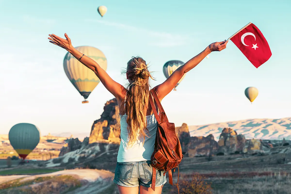 una mujer vestida con camiseta y pantalones cortos y situada de espaldas ondea una bandera turca mientras contempla alzarse los globos aerostáticos sobre el paisaje de Capadocia