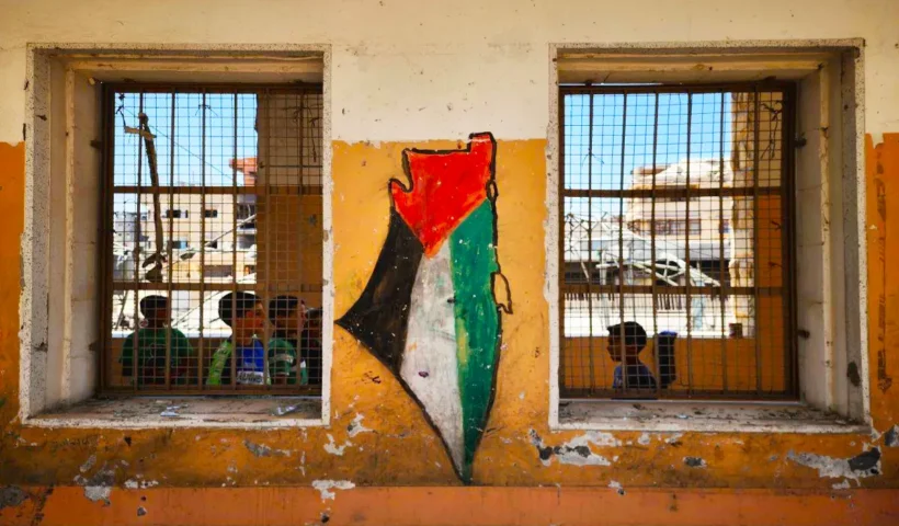 unos niños palestinos en el interior de una casa en ruinas con verjas en Cisjordania sobre cuya fachada hay pintada una bandera de Palestina
