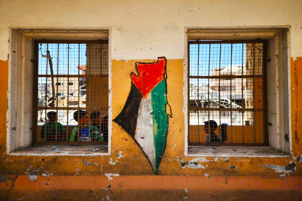 unos niños palestinos en el interior de una casa en ruinas con verjas en Cisjordania sobre cuya fachada hay pintada una bandera de Palestina