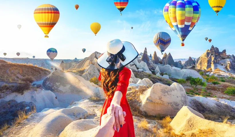 una mujer sosteniendo la mano de un hombre mientras lo conduce hacia un paisaje de la Capadocia rodeado de globos aerostáticos volando