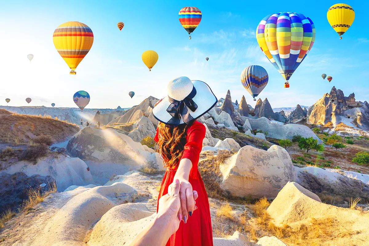 una mujer sosteniendo la mano de un hombre mientras lo conduce hacia un paisaje de la Capadocia rodeado de globos aerostáticos volando