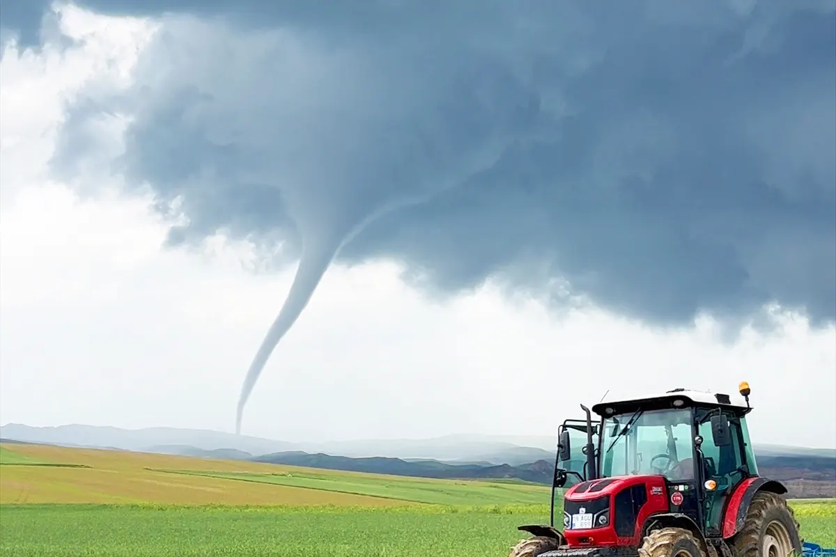 an abandoned tractor in a green field, with a huge tornado forming a short distance away, emerging from a dense grey cloud
