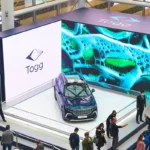 varias personas en torno a un gran stand en una feria donde se exhibe rodeado de grandes pantallas una unidad del coche eléctrico turco TOGG