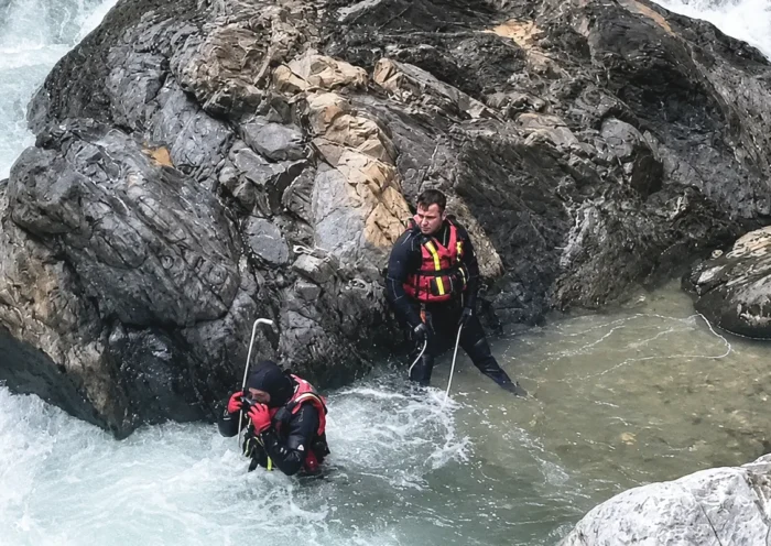 Two search-and-rescue personnel are searching a fast-flowing river amid large boulders and water