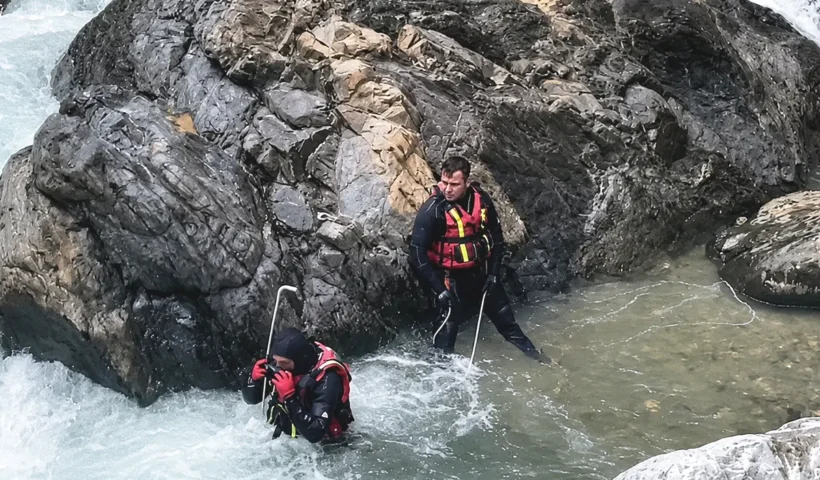 Two search-and-rescue personnel are searching a fast-flowing river amid large boulders and water
