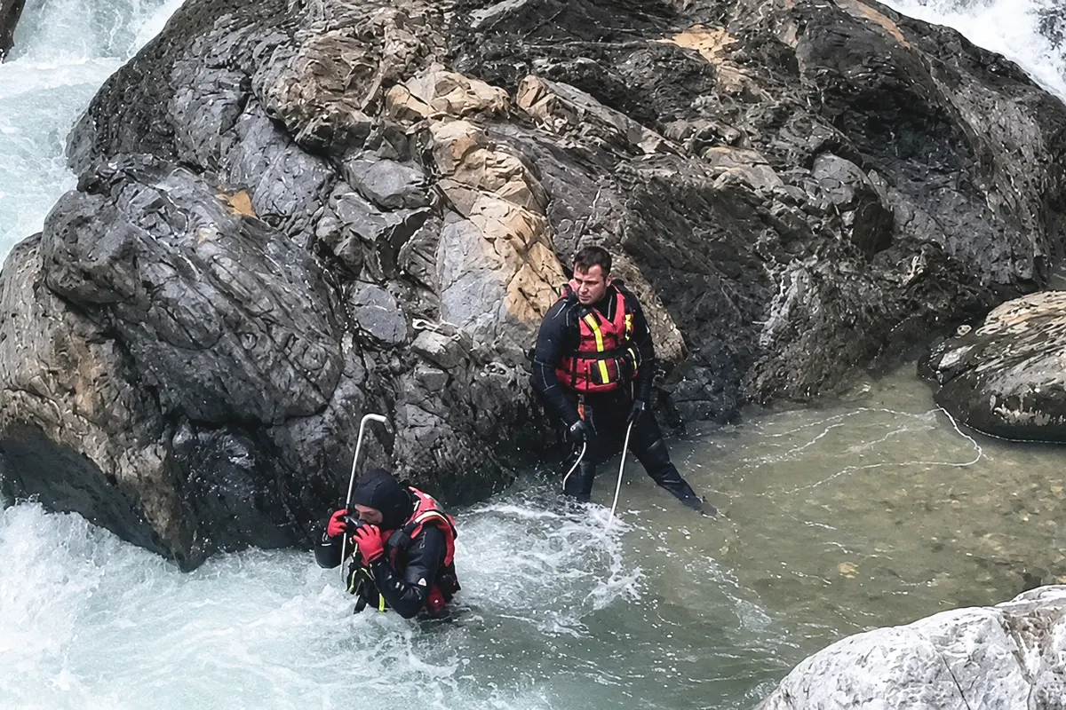 Two search-and-rescue personnel are searching a fast-flowing river amid large boulders and water