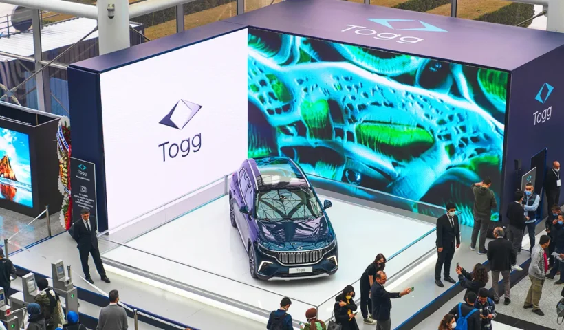 Several people gathered around a large booth at a trade show where a Turkish TOGG electric car is on display, surrounded by large screens.