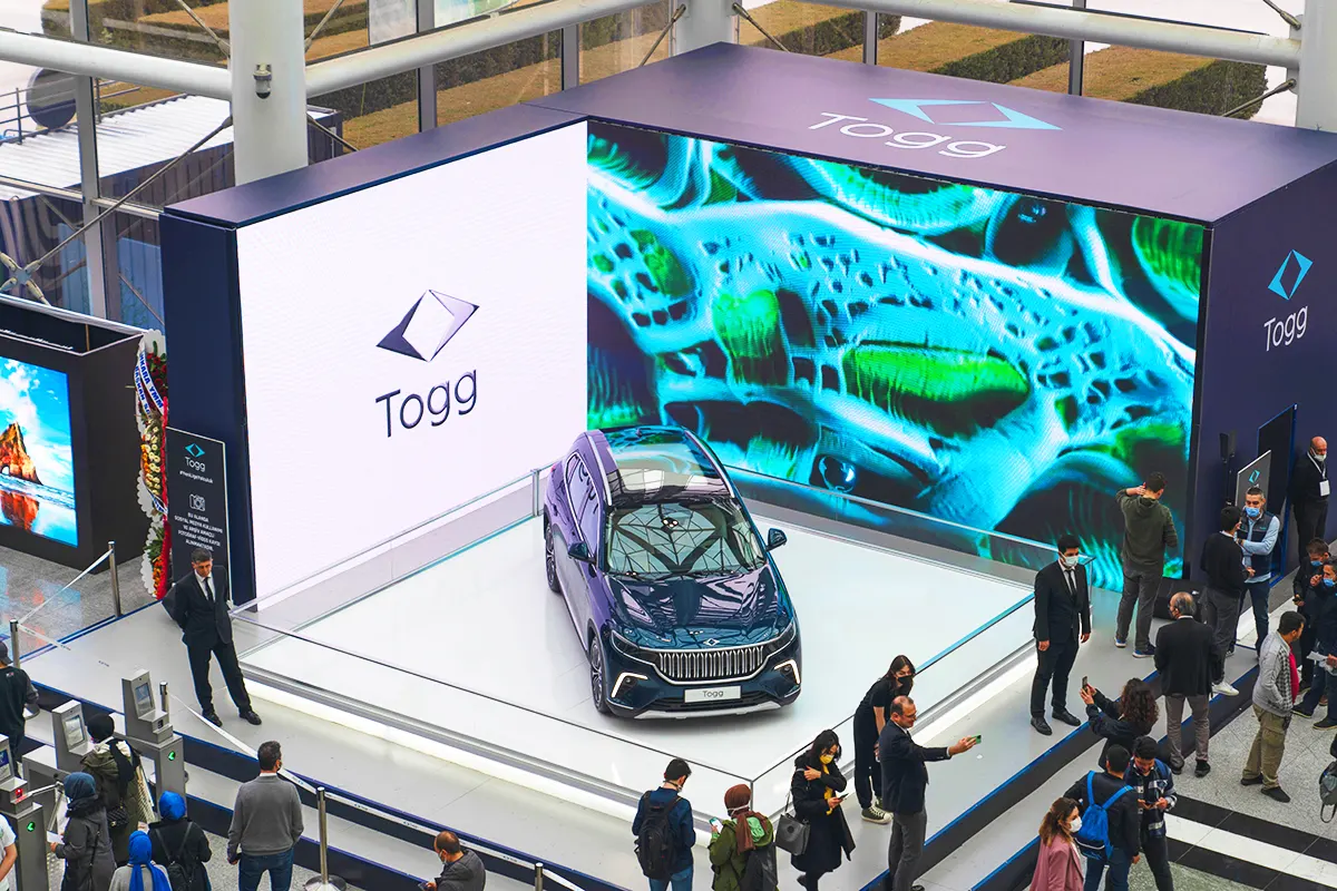 Several people gathered around a large booth at a trade show where a Turkish TOGG electric car is on display, surrounded by large screens.