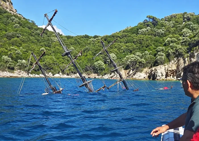 Several people are looking at the masts of a sunken ship off the coast, protruding from the water