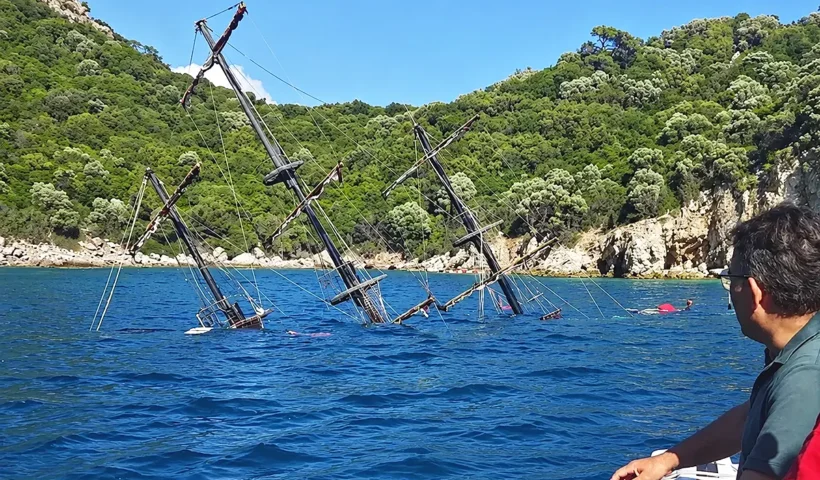 Several people are looking at the masts of a sunken ship off the coast, protruding from the water