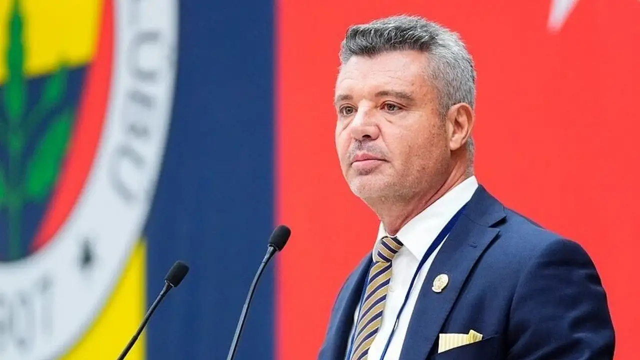 el presidente del Fenerbahçe, Sadettin Saran, aparece en primer plano hablando por dos micrófonos; de fondo se ve un enorme escudo del Fenerbahçe