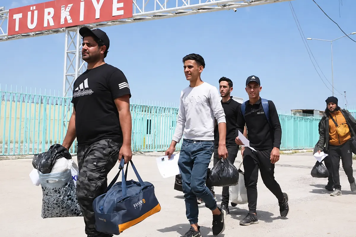 un grupo de inmigrantes atraviesa la frontera cargado con bolsas bajo un cartel que pone "Türkiye"