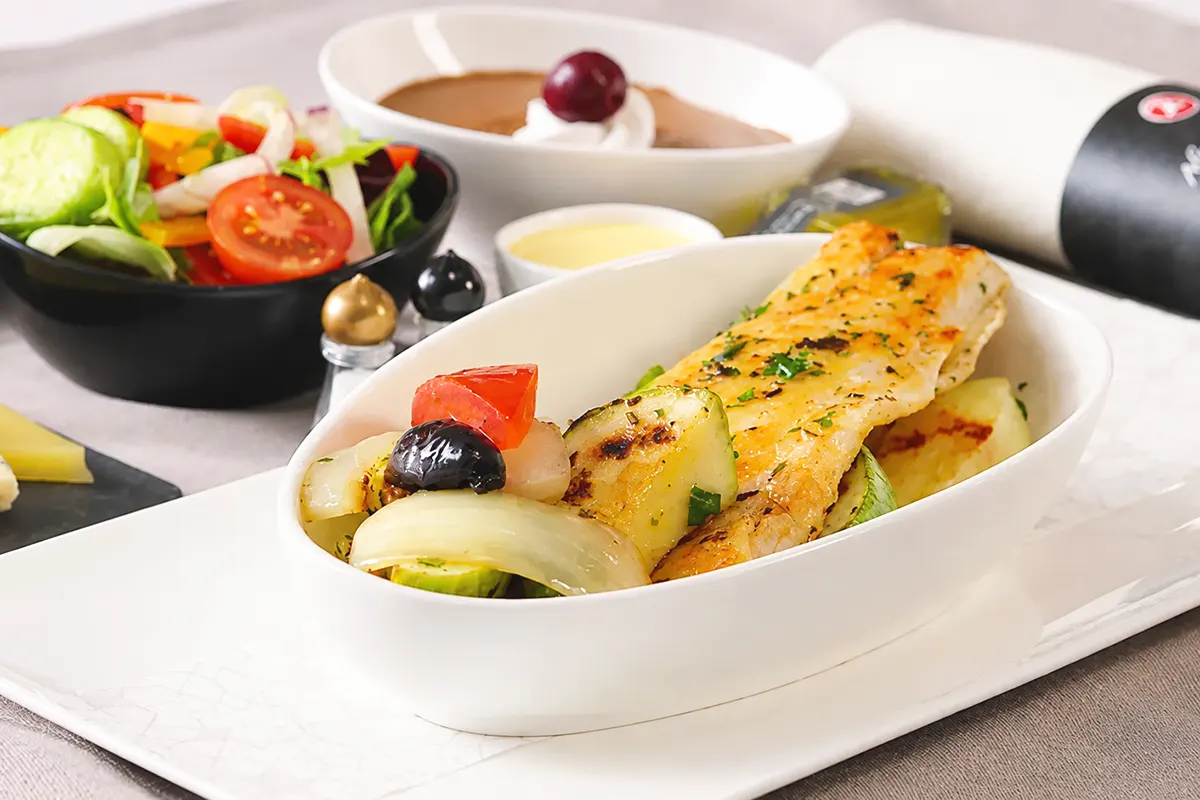 un menú de avión de Turkish Airlines mostrando en primer plano un plato de pescado junto a un cuenco con ensalada, un postre de crema y una servilleta enrollada