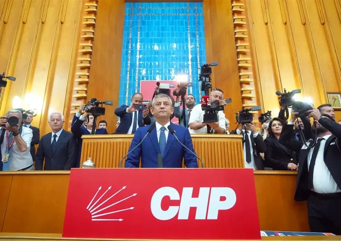 Özgür Özel standing on a podium adorned with a large red banner bearing the initials of the CHP party, surrounded by journalists