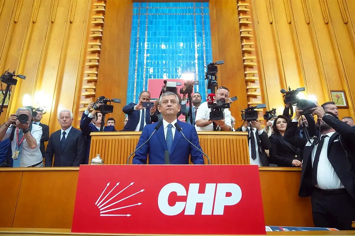 Özgür Özel standing on a podium adorned with a large red banner bearing the initials of the CHP party, surrounded by journalists