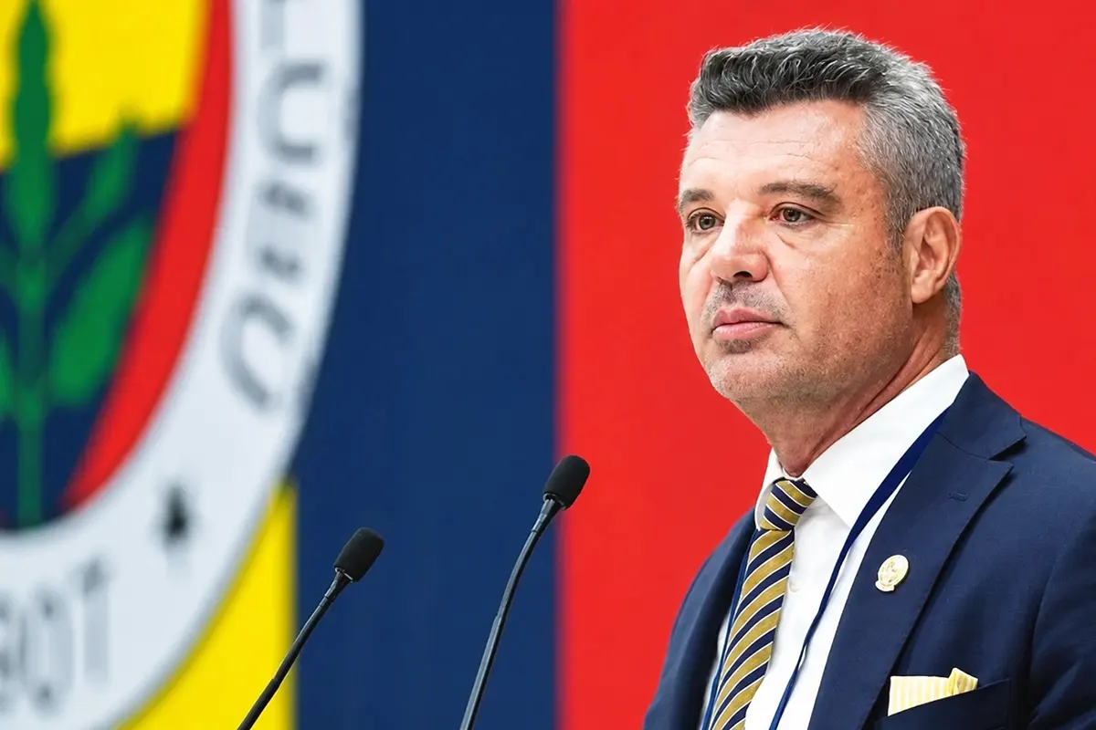 Fenerbahçe President Sadettin Saran is shown in the foreground speaking into two microphones; in the background is a huge Fenerbahçe crest