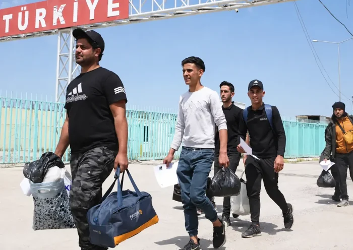 A group of migrants crosses the border, laden with bags, beneath a sign reading ‘Türkiye’