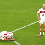a Turkish player standing on the pitch looking stunned, and another beside him on his knees with his face pressed against the grass, crying