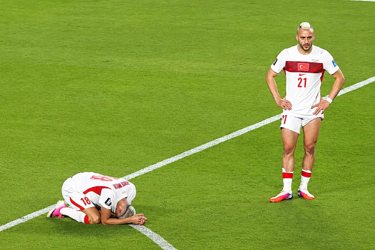 a Turkish player standing on the pitch looking stunned, and another beside him on his knees with his face pressed against the grass, crying