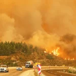 several vehicles travelling along a motorway next to a burning hill and surrounded by smoke, set against an orange, almost apocalyptic backdrop