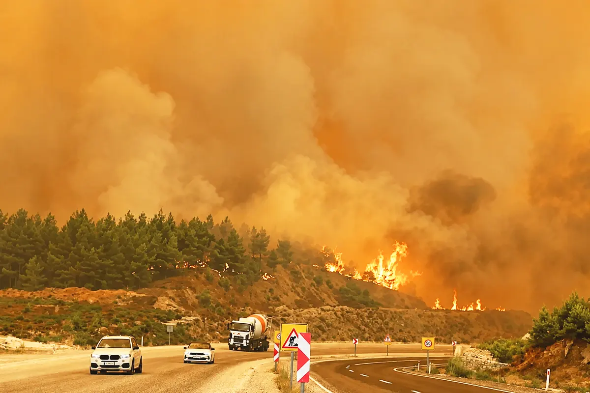 varios vehículos circulando por una autopista junto a un monte ardiendo y rodeado de humo, en medio de un paisaje anaranjado y cuasi-apocalíptico