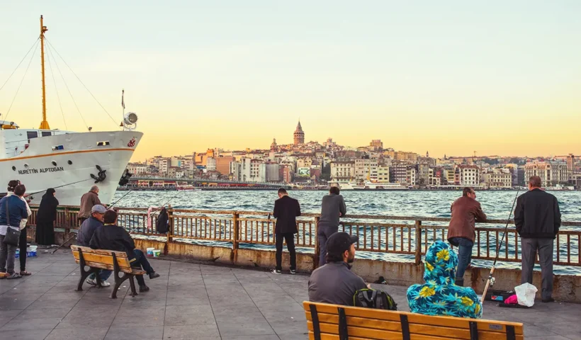Several people sitting and looking out over the waters of the Golden Horn in Istanbul, next to a ferry; the Galata Tower can be seen in the background