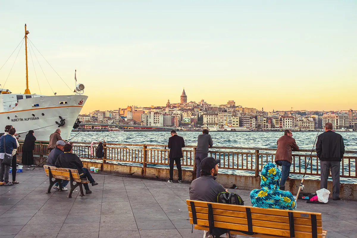 Several people sitting and looking out over the waters of the Golden Horn in Istanbul, next to a ferry; the Galata Tower can be seen in the background