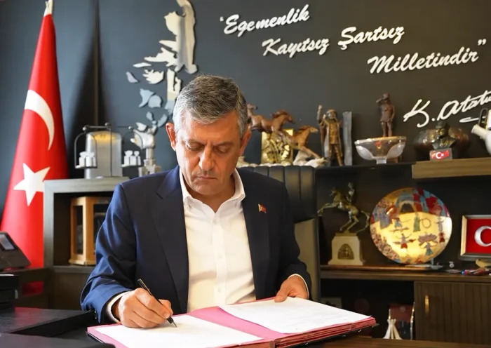 Özgür Özel firma unos documentos en un despacho; de fondo hay una bandera turca, varios objetos decorativos, y una pared con una frase firmada por Atatürk