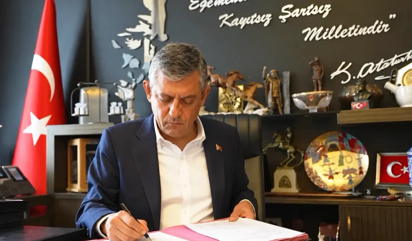 Özgür Özel firma unos documentos en un despacho; de fondo hay una bandera turca, varios objetos decorativos, y una pared con una frase firmada por Atatürk