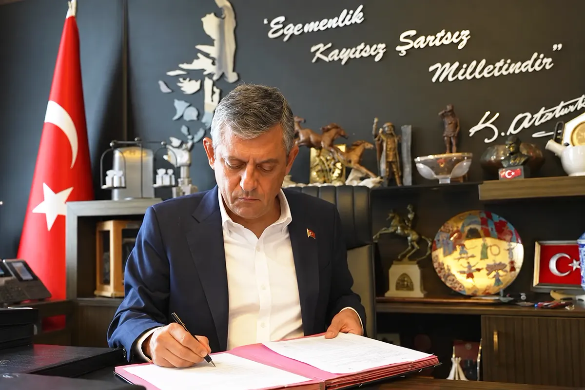 Özgür Özel firma unos documentos en un despacho; de fondo hay una bandera turca, varios objetos decorativos, y una pared con una frase firmada por Atatürk