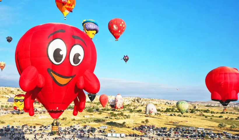 decenas de globos de varias formas y colores ascienden sobre el paisaje de Capadocia, en primer plano aparece uno de color rojo que representa a un simpático cangrejo sonriente