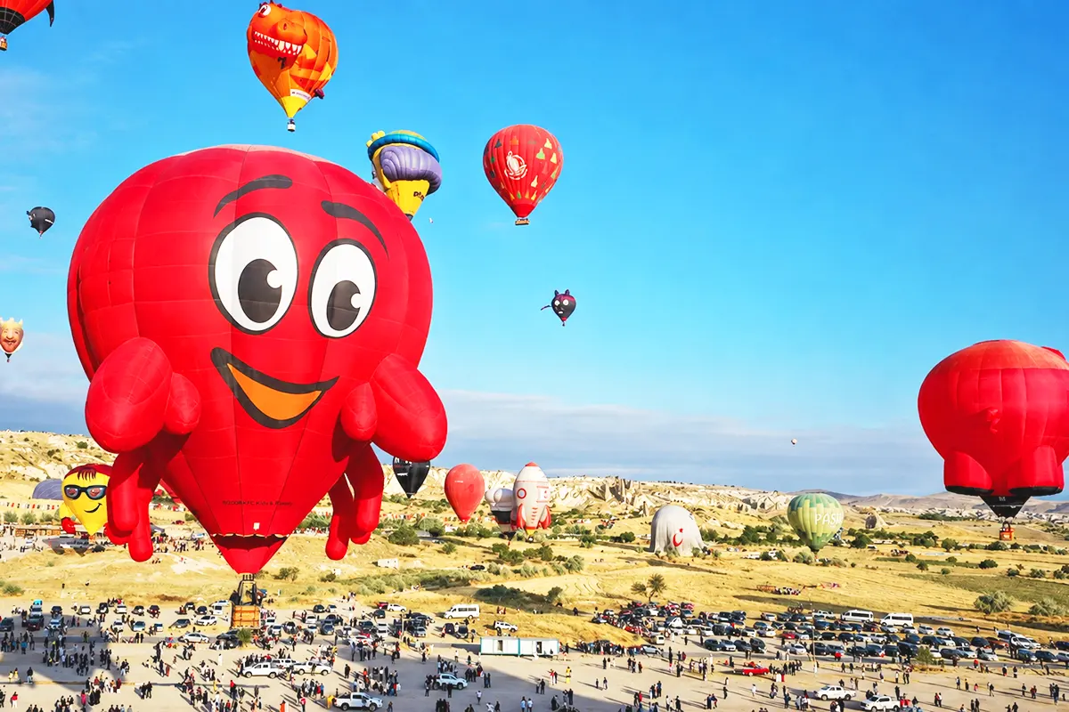 decenas de globos de varias formas y colores ascienden sobre el paisaje de Capadocia, en primer plano aparece uno de color rojo que representa a un simpático cangrejo sonriente