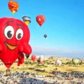 Dozens of hot-air balloons of various shapes and colours rise above the landscape of Cappadocia; in the foreground is a red one depicting a friendly, smiling crab