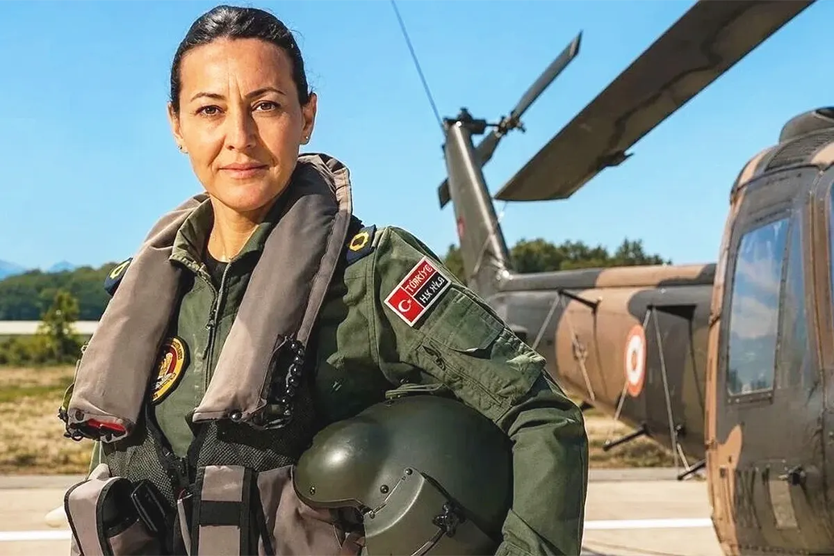 A woman looking at the camera, wearing a Turkish Air Force pilot’s uniform, poses with a helmet under her arm next to a helicopter